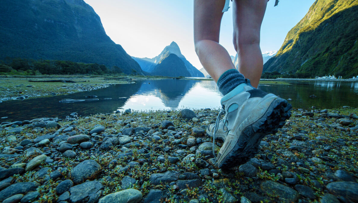 Zwei Beine einer Person in Wanderschuhen an einem See vor Bergpanorama.