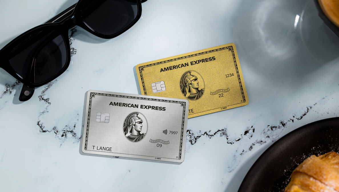 Eine silberne und eine goldene Kreditkarte von American Express liegen auf einem weißen Marmortisch neben einer schwarzen Sonnenbrille, einer Kaffeetasse und einem Teller mit Gebäck.