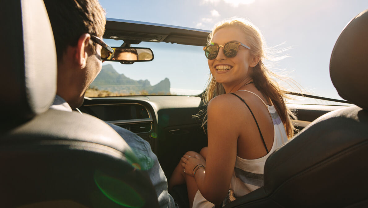 Eine junge Frau mit Sonnenbrille auf dem Beifahrersitz eines Cabriolets schaut lachend zu ihrem Partner hinüber, im Hintergrund eine Küstenlandschaft bei Sonnenschein.