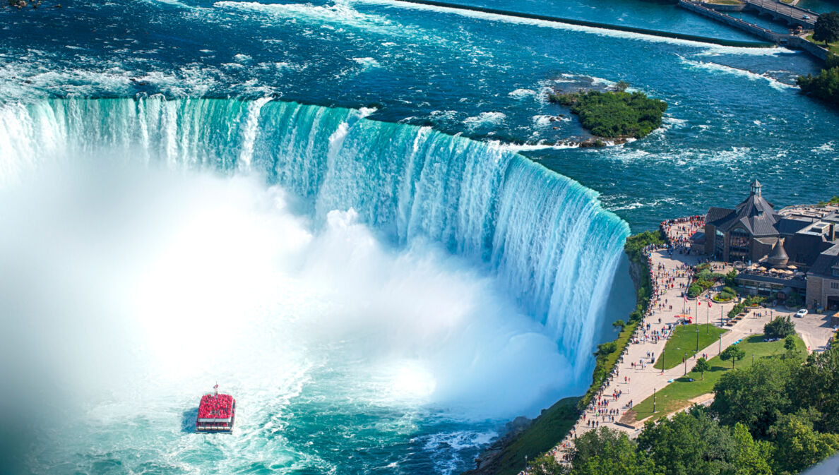 Luftaufnahme eines runden Wasserbeckens mit Boot vor einem Wasserfall, am Ufer eine Aussichtsplattform mit Personen.