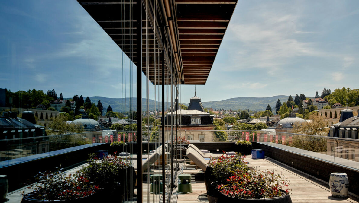 Große Balkonterrasse mit Liegestühlen vor grüner Hügellandschaft, die sich in der Glasfassade des Gebäudes spiegelt.