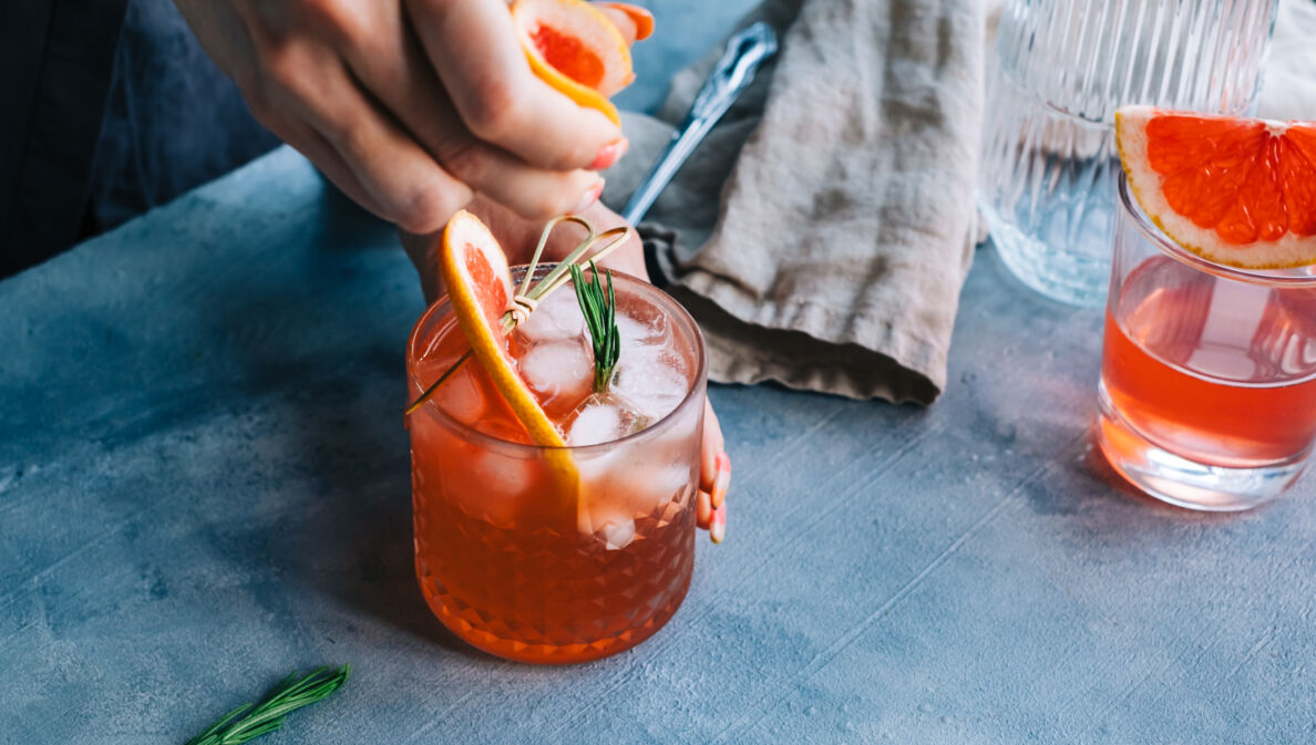 Eine Nahaufnahme einer Person, die einen Cocktail anrichtet, der mit einer Grapefruit-Scheibe und Rosmarin verziert ist.