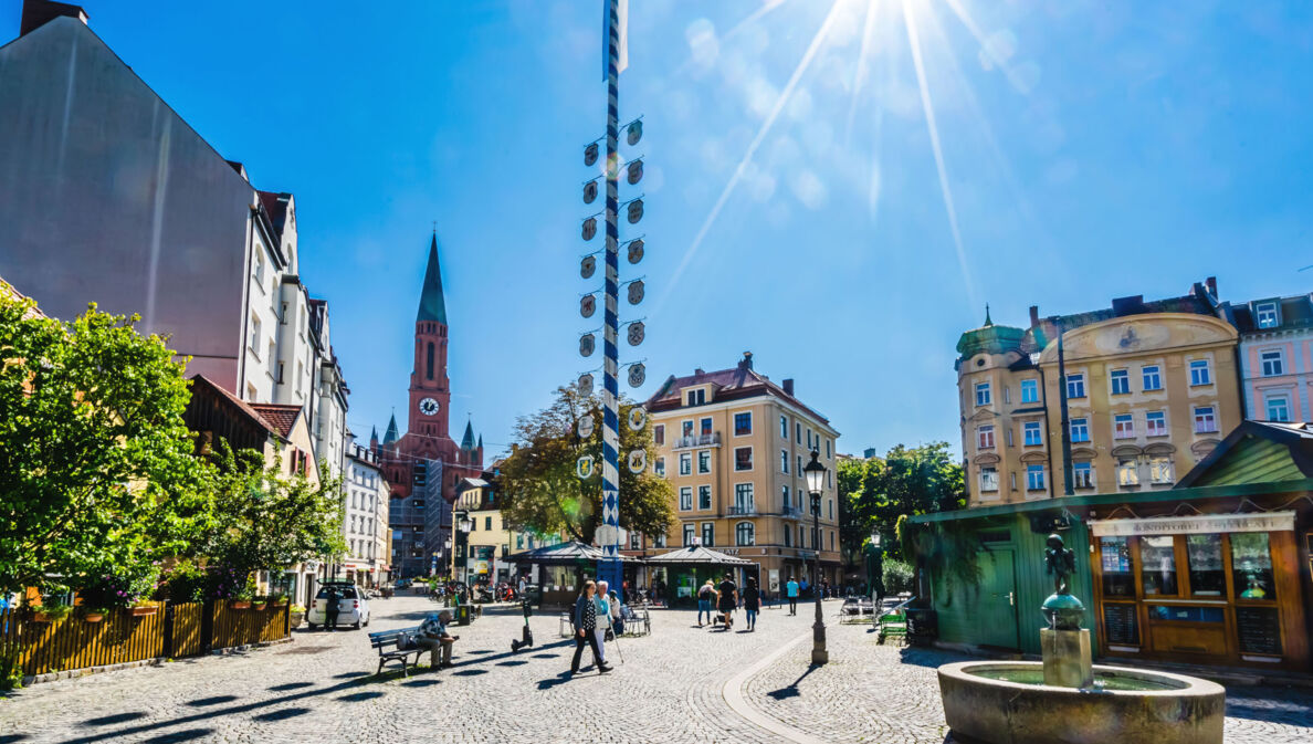 Ein Stadtteilplatz mit Kopfsteinpflaster und einem Maibaum in der Mitte bei strahlendem Sonnenschein.