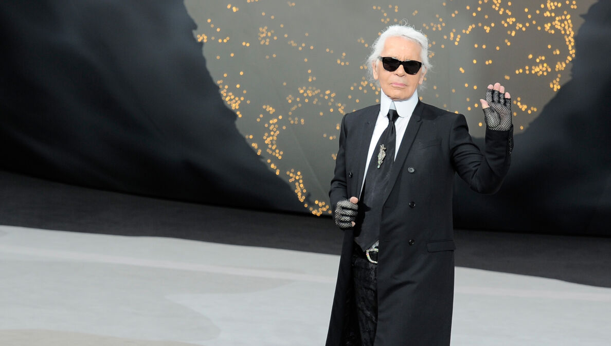 Karl Lagerfeld mit Sonnenbrille im schwarzen Jackett winkt vom Laufsteg einer Modenschau.