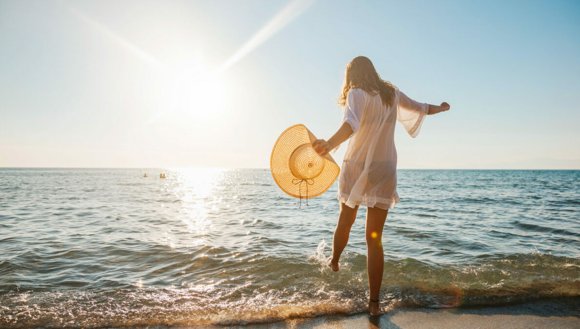 Eine junge Frau im weißen Sommerkleid und mit Strohhut in der Hand planscht mit ihrem Fuß im Wasser an einem Sandstrand am Meer bei Sonnenuntergang