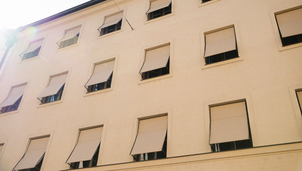 Hellbeige Hausfassade mit gleichfarbigen Markisen vor den Fenstern.