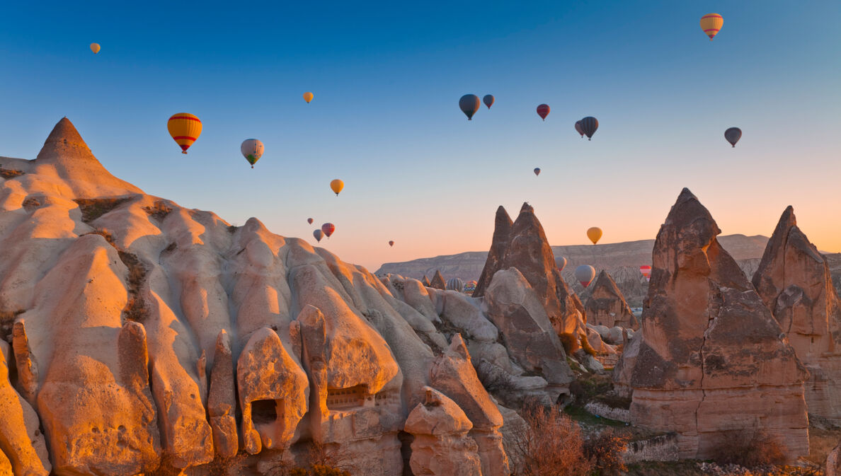 Felsige Landschaft in Kappadokien bei Sonnenuntergang mit zahlreichen schwebenden Heißluftballons im Hintergrund.