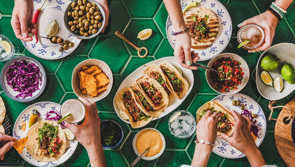 Aufnahme eines Tisches von oben, an dem mehrere Personen sitzen. Auf dem Tisch stehen verschiedene Teller mit mexikanischem Essen wie Tacos, Dips, Nachos und mehr.