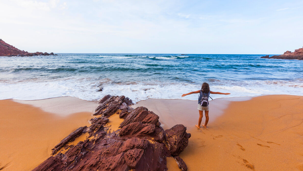 Eine Person steht mit dem Rücken zur Kamera und neben einem Felsen an einem Strand und schaut mit ausgebreiteten Armen aufs Meer