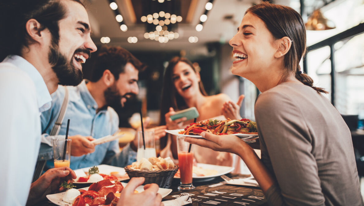 Eine Gruppe fröhlicher Personen in einem Restaurant. Eine lachende Frau hält einen Teller in der Hand.