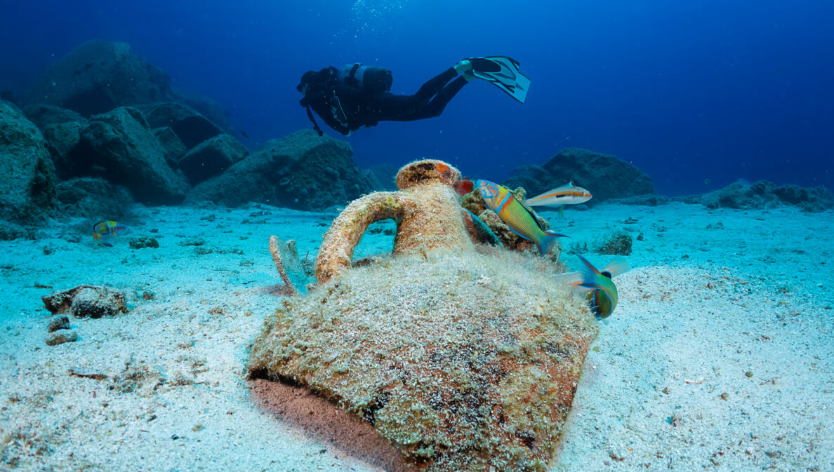 Eine zerbrochene Amphore auf dem Meeresgrund, die von bunten Fischen umgeben ist. Im Hintergrund schwimmt eine Person in Tauchausrüstung.