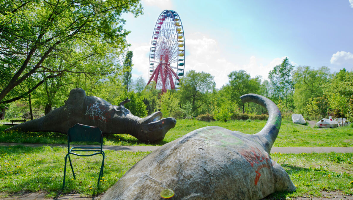 Zwei umgekippte Dinosaurier-Skulpturen auf einer Wiese vor einem Riesenrad in einem Freizeitpark.