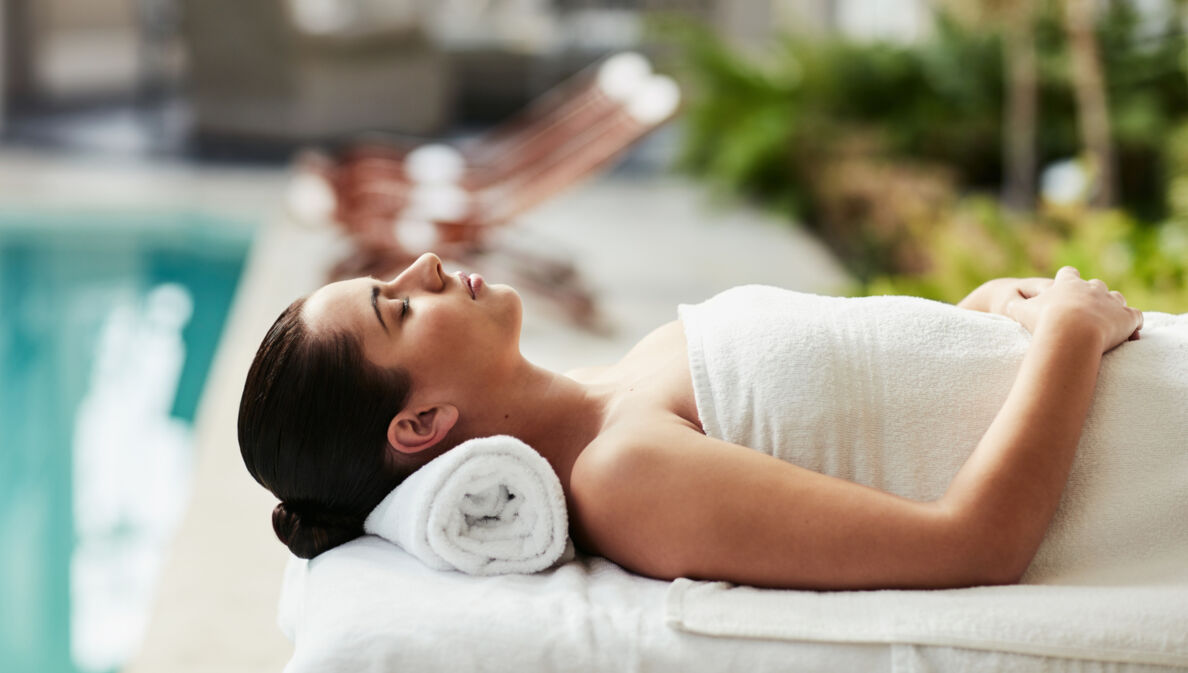 Eine Frau entspannt in einem weißen Handtuch auf einer Massageliege an einem Hotelpool.