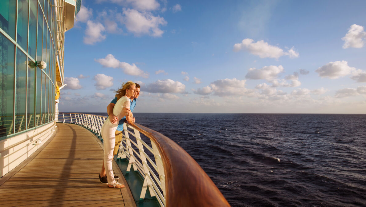 Ein Paar steht an der Reling eines Kreuzfahrtschiffes und schaut aufs Meer im Sonnenschein.