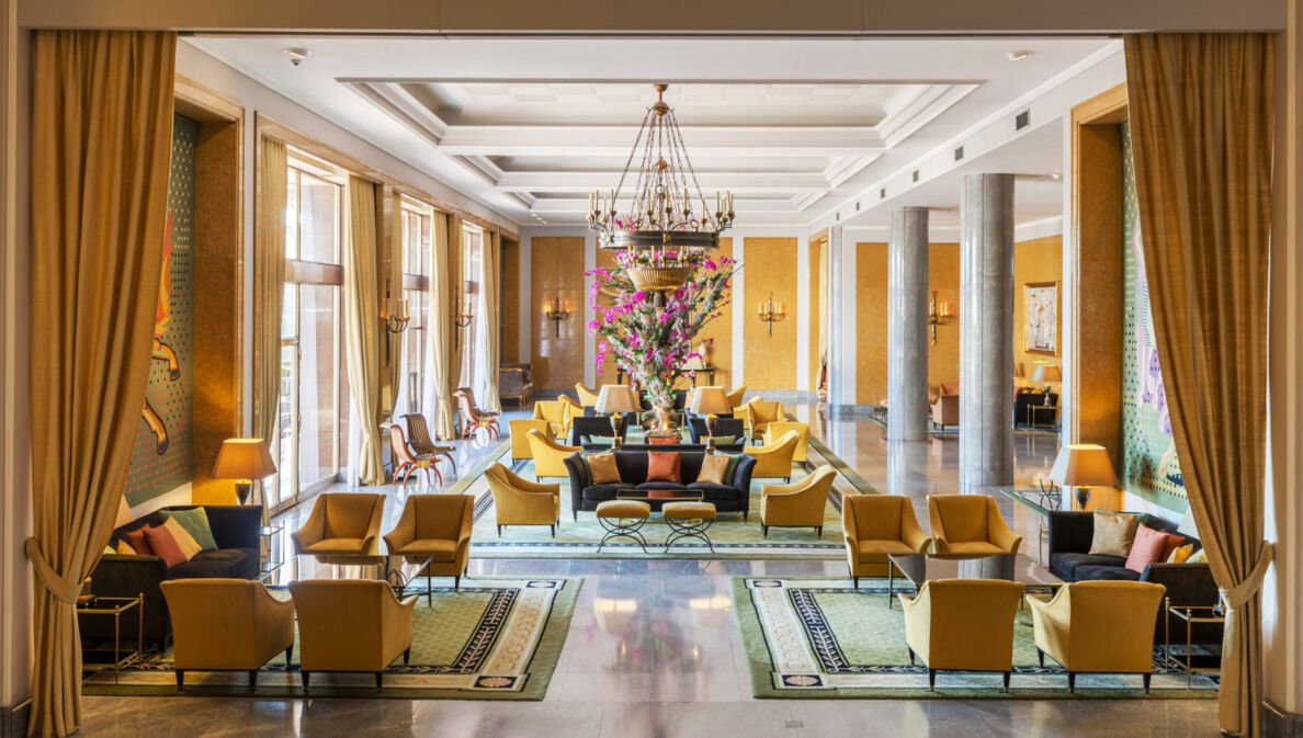 Ein elegantes Hotelfoyer mit Marmorsäulen und gelben Sesseln, in der Mitte ein großes Blumenbouquet.