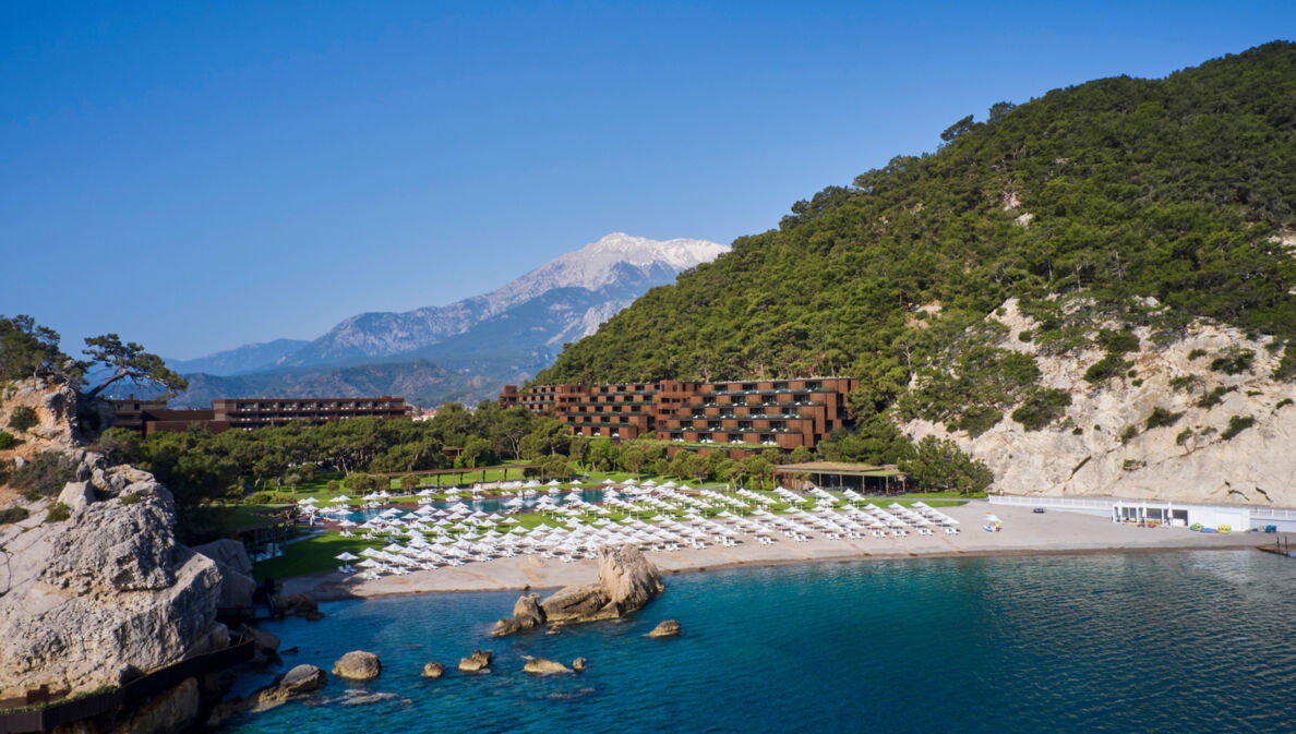 Ein Hotelresort in einer mediterranen Bucht mit Sandstrand, im Hintergrund eine Berglandschaft.