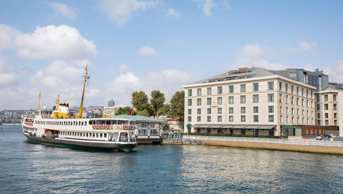 Gebäude mit neoklassizistischer Fassade am Ufer eines Gewässers mit Schiff im Vordergrund, im Hintergrund die Skyline von Istanbul.