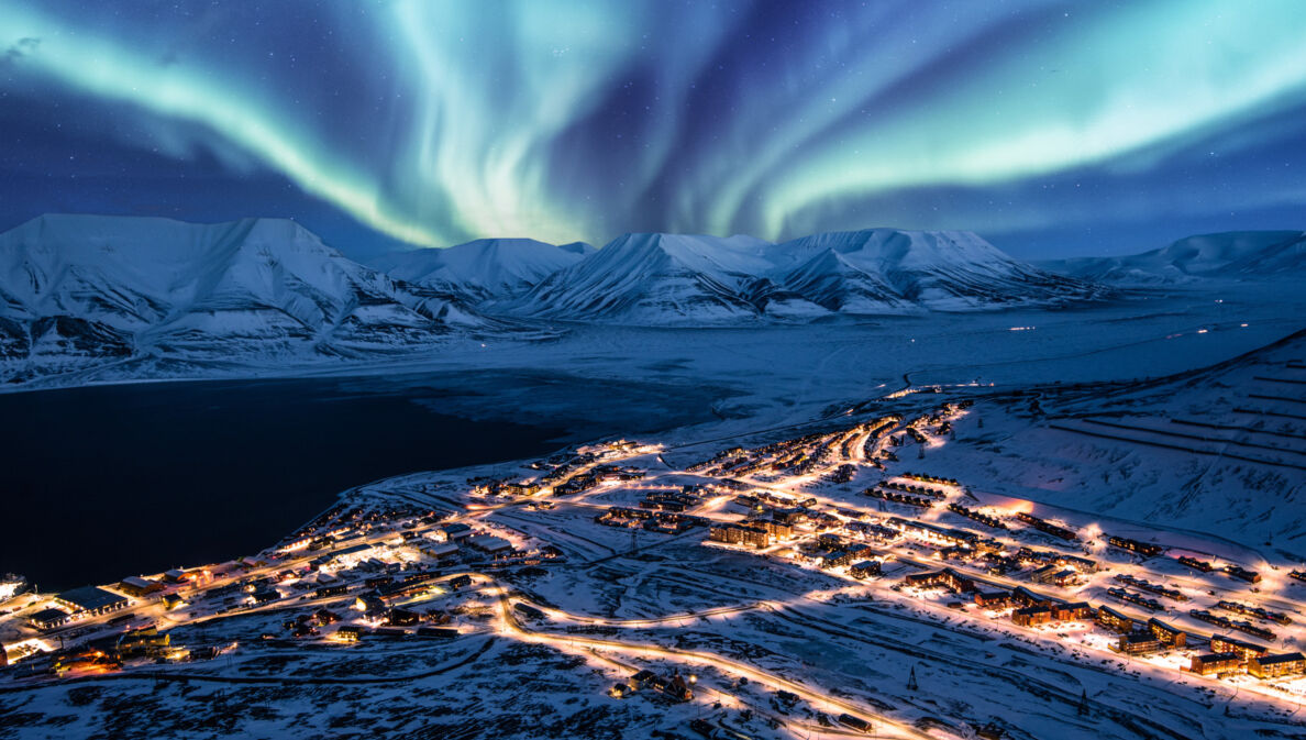 Beleuchtete Ortschaft in schneebedeckter Fjordlandschaft unter Polarlichtern bei Nacht.