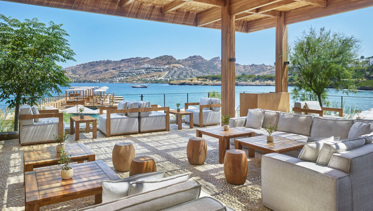 Loungebereich mit grauen Sesseln und Holztischen auf einer überdachten Terrasse an einer mediterranen Bucht.