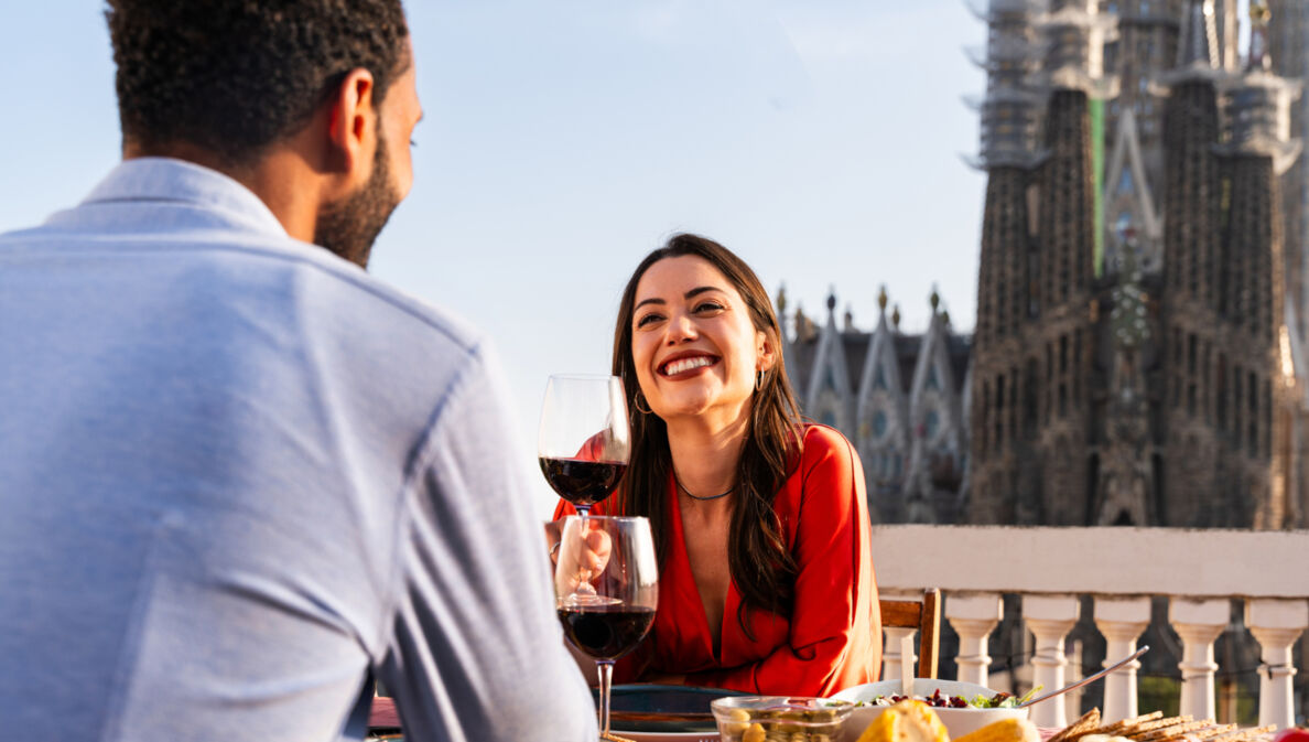 Eine Frau sitzt gegenüber von einem Mann an einem Tisch und hält lächelnd ein Glas Rotwein in der Hnad. Im Hintergrund die Sagrada Familia von Barcelona.