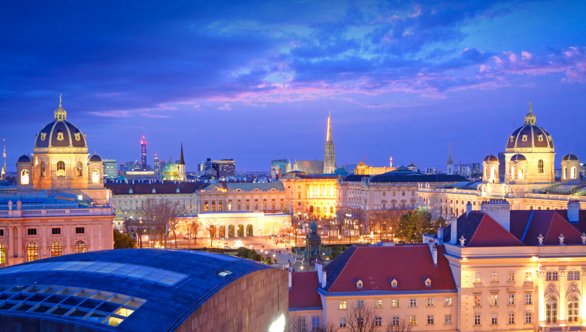 Blick von oben auf eine Stadt in der Dämmerung. Im Vordergrund sind prächtige, beleuchtete Gebäude zu sehen.