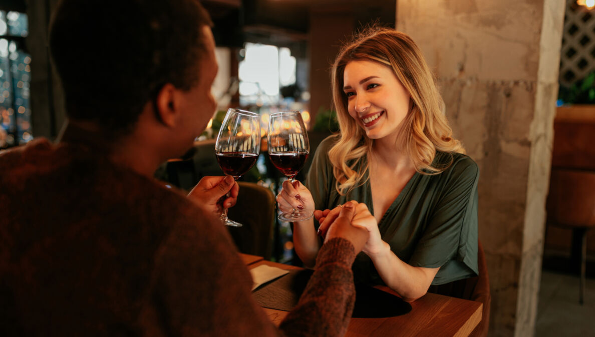 Eine lächelnde Frau sitzt einem Mann an einem Restauranttisch gegenüber und hält seine Hand. In der anderen Hand halten sie je ein Glas Rotwein.