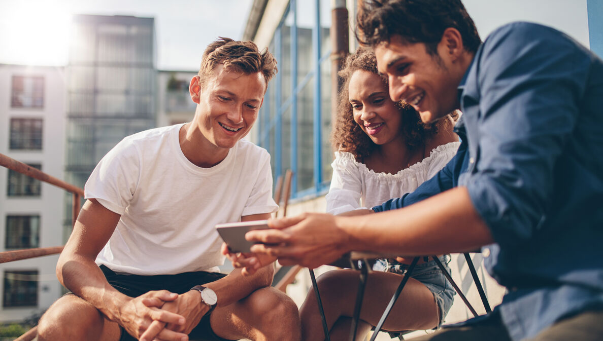 Eine Gruppe von Menschen sitzt zusammen auf dem Balkon und schaut gemeinsam etwas auf dem Smartphone.