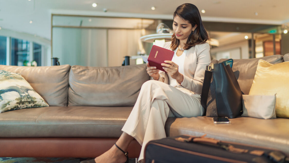 Eine Frau sitzt mit überschlagenen Beinen auf einem hellen Ledersofa und sieht sich Reiseunterlagen an.