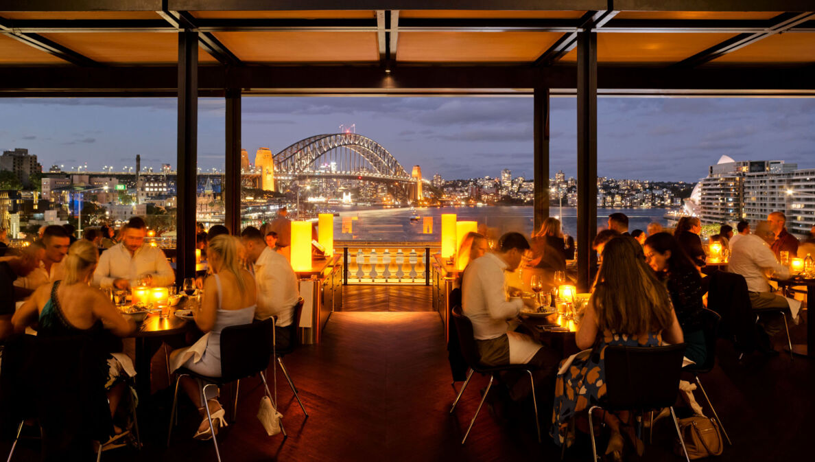 Außengastronomie am Hafen von Sydney vor beleuchteter Harbour Bridge bei Nacht.