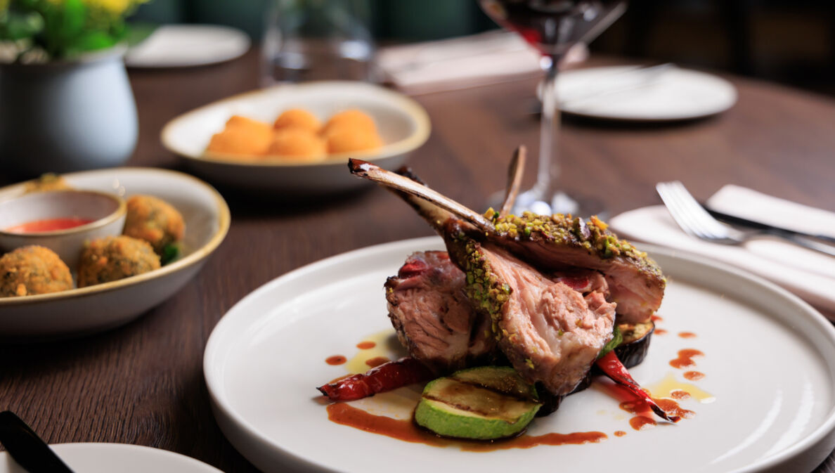 Gegrilltes Lammrippenkotelett, angerichtet auf einem weißen Teller, daneben Beilagen auf einem Tisch in einem Restaurant.