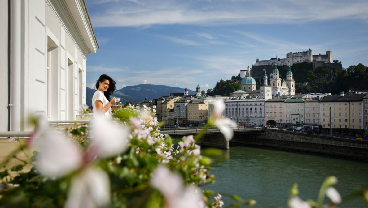 Eine Frau steht mit Kaffeetasse auf einem Balkon am Fluss, ihr gegenüber die Salzburger Altstadt mit Burgfestung.