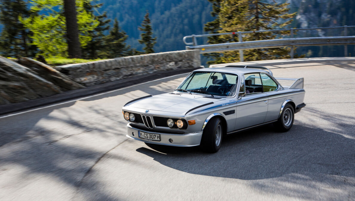 Ein fahrender silberner BMW 3.0 CSL in einer Kurve auf einer Straße in den Bergen.