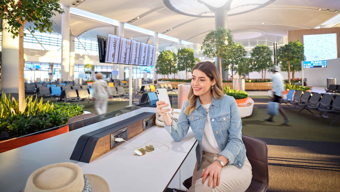 Eine junge Frau mit Smartphone sitzt in einem Wartebereich am Flughafen.