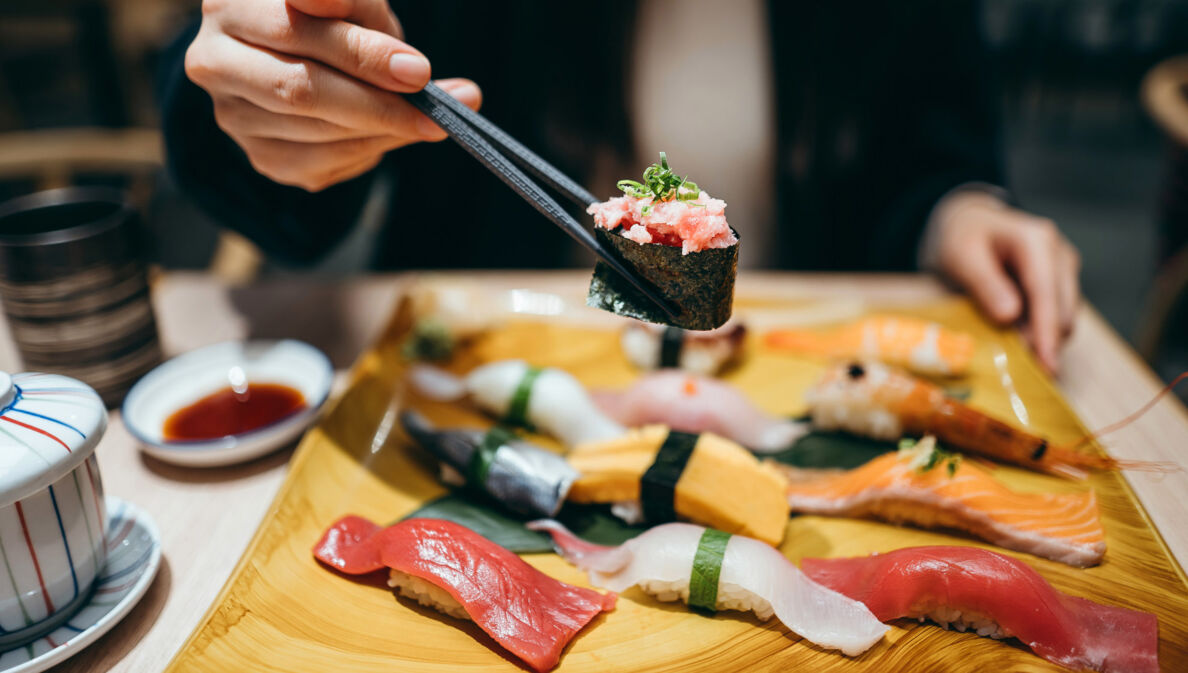 Eine Person hält eine Rolle Maki Sushi zwischen zwei Essstäbchen über einer Bambusplatte mit Nigiri auf einem Tisch in einem Restaurant.