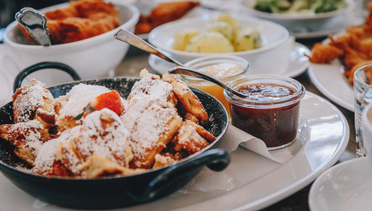 Ein schwarzer Topf mit Kaiserschmarrn auf einem weißen Teller, drumherum kleine Schälchen mit verschiedenen Leckereien.