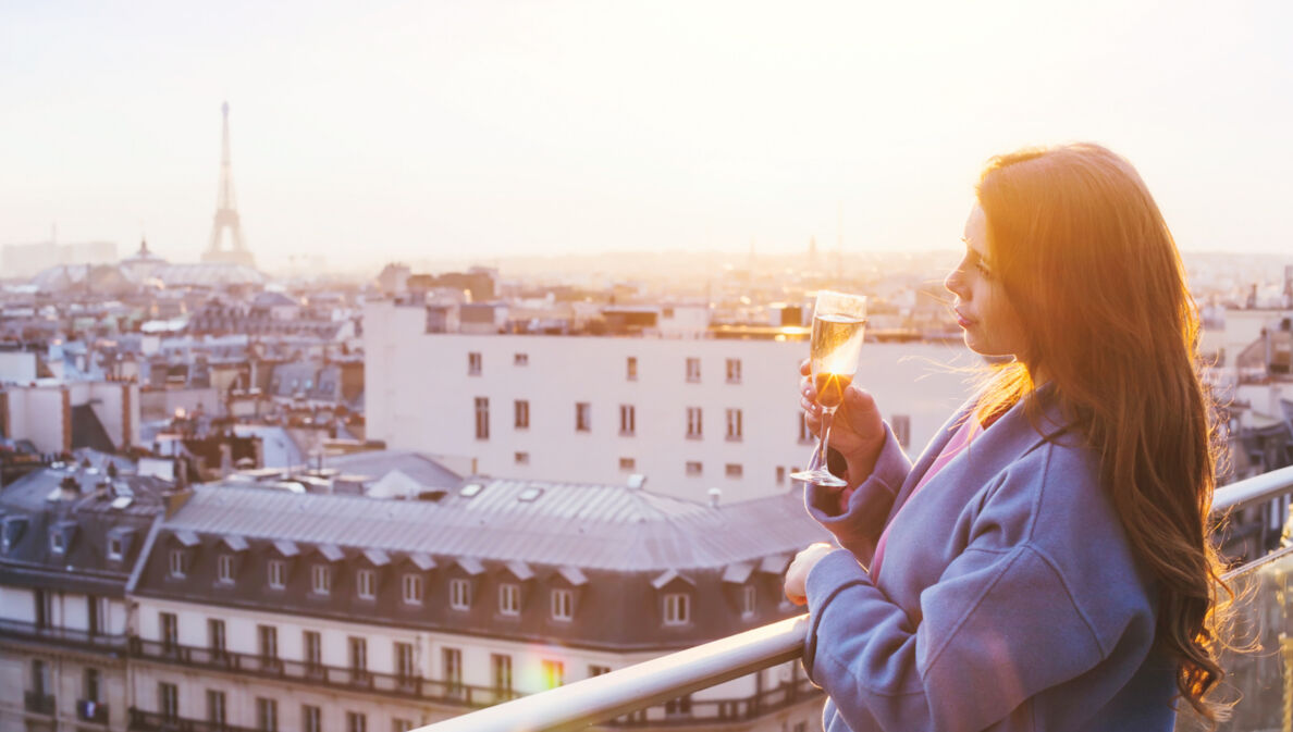 Eine Frau, die bei Sonnenuntergang in Paris auf einem Balkon steht und ein Glas Sekt in der Hand hält.