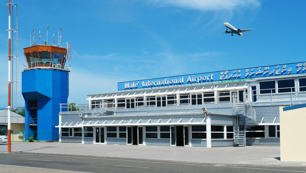 Ein Flugzeug fliegt am blauen Himmel über einen kleines Flughafengebäude mit blauem Kontrollturm.