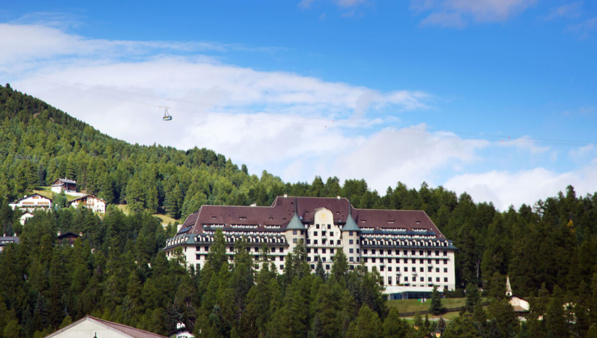 Ein Grandhotel mit zwei Türmen in einem Waldgebiet auf einem Berg, darüber fährt eine Seilbahn.