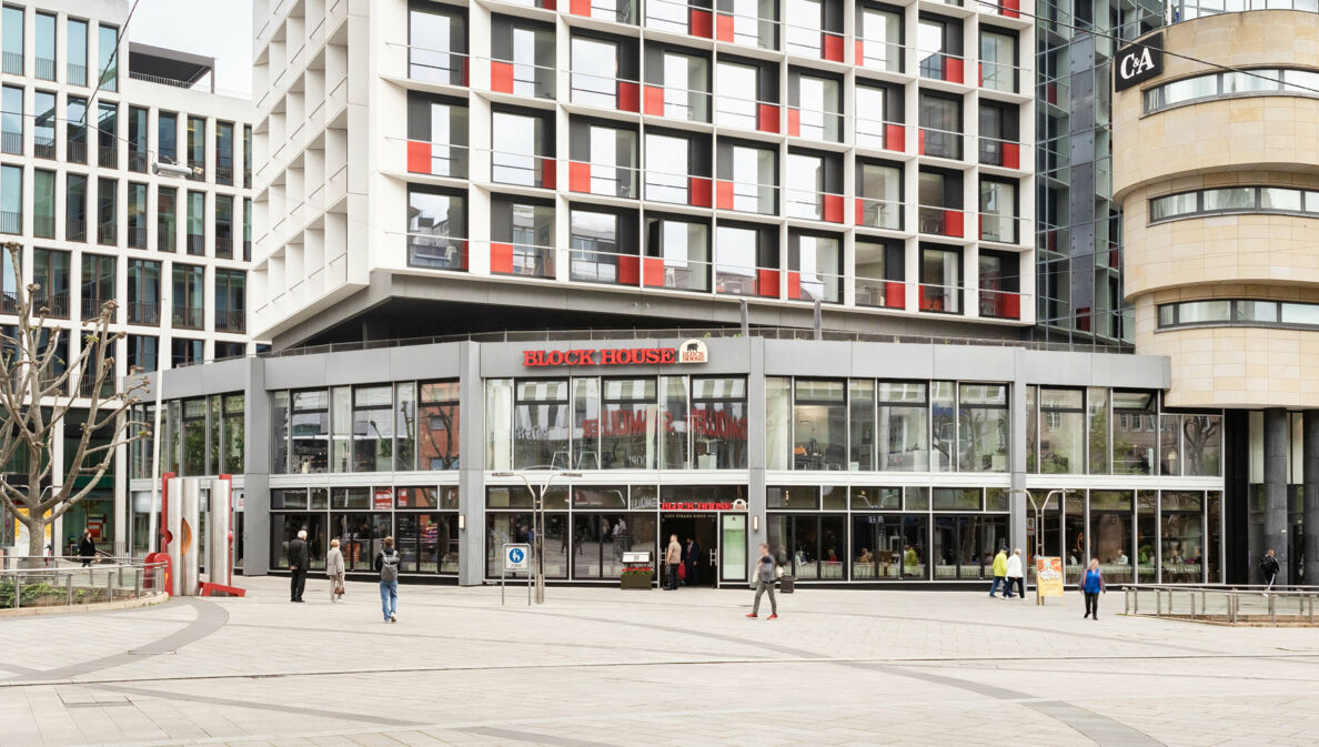 Personen am Eingang einer Block House Filiale in einem grauen Neubau in einer Fußgängerzone in Innenstadtlage.