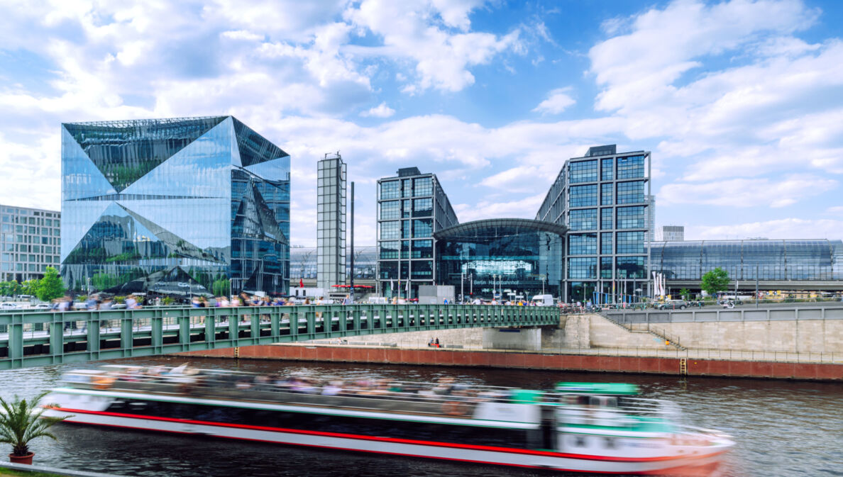 Modernes Bauensemble des Berliner Hauptbahnhofes mit Glasfassade am Flussufer, im Vordergrund ein Ausflugsboot in Unschärfe.