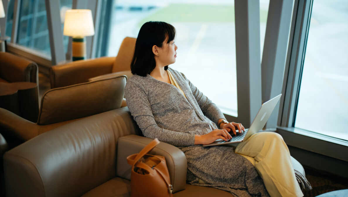 Eine Frau sitzt mit Laptop in einem Ledersessel am Fenster einer Flughafen-Lounge und schaut aufs Rollfeld.