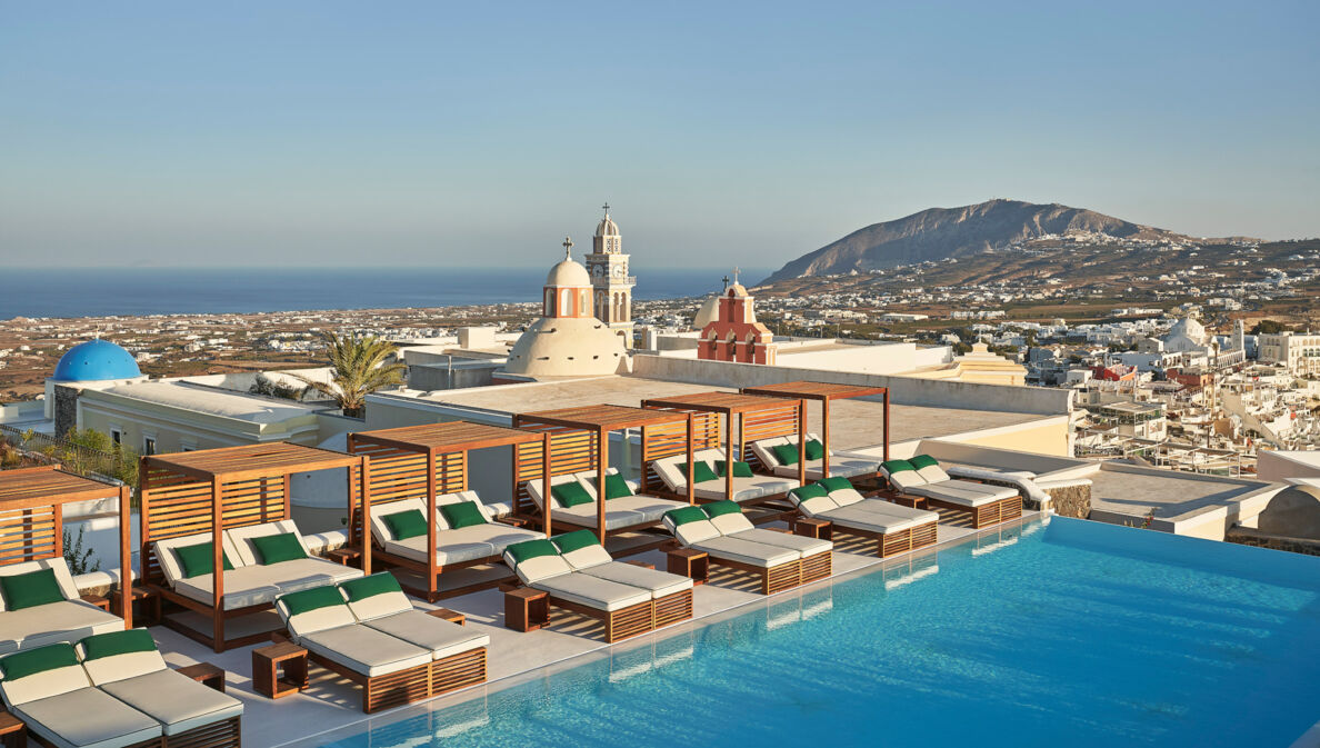 Dachterrasse mit zahlreichen Sonnenliegen am Infinity-Pool auf Santorini, im Hintergrund Meeresküste.