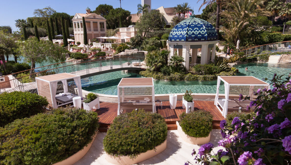 Ein lang gestreckter, mit Palmen, Blumen und Daybeds gesäumter Pool, über den zwei Brücken führen.