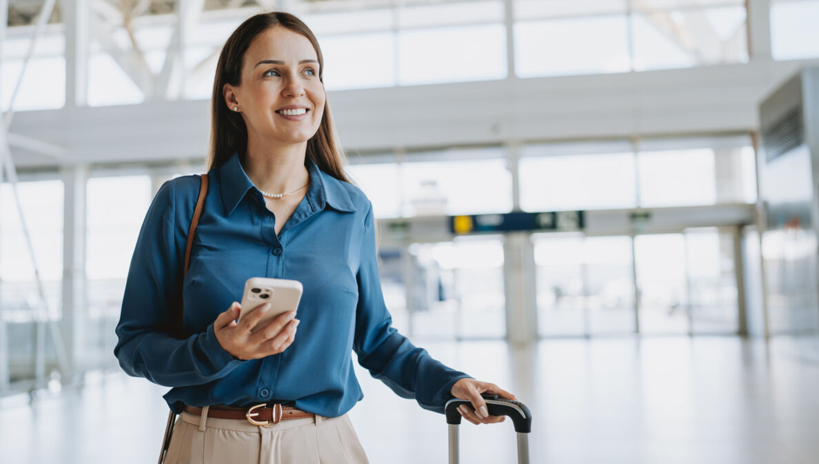 Eine Frau am Flughafen, die in einer Hand einen Koffergriff hält und in der anderen ein Smartphone.