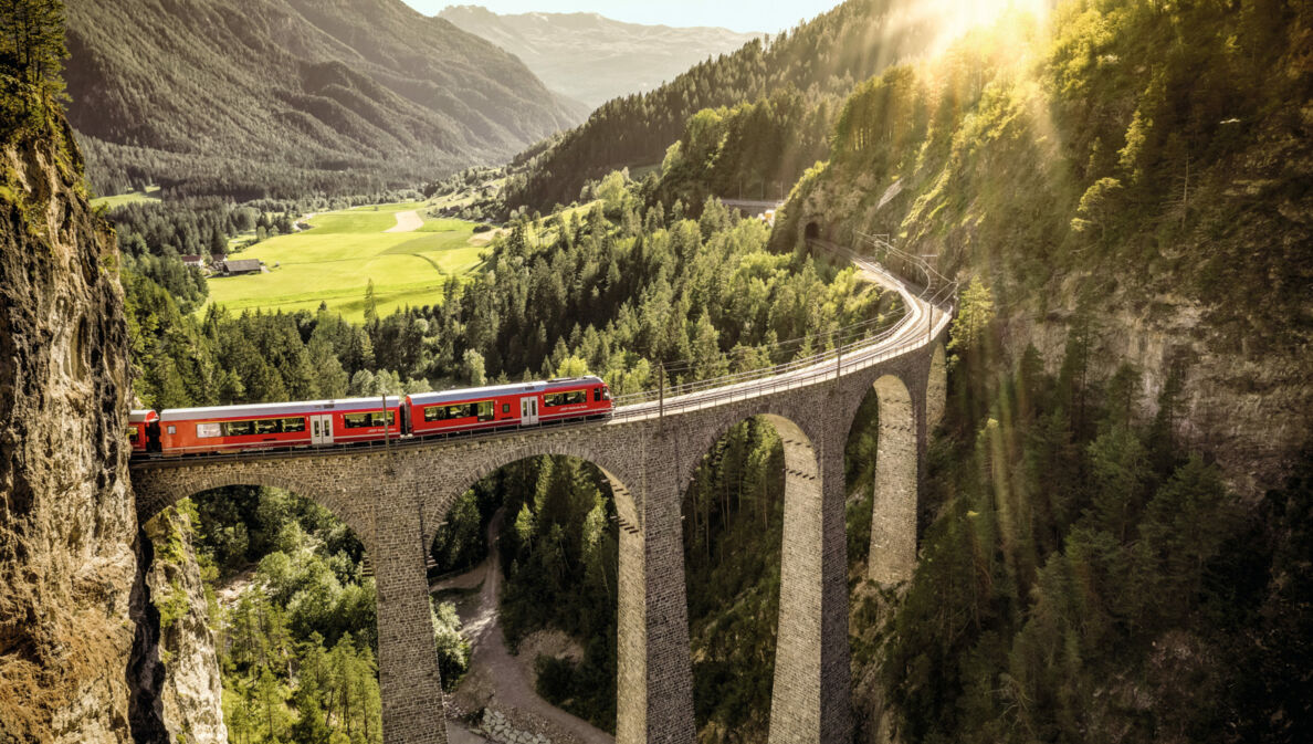 Ein roter Zug fährt auf einem Viadukt durch eine grüne Berglandschaft.