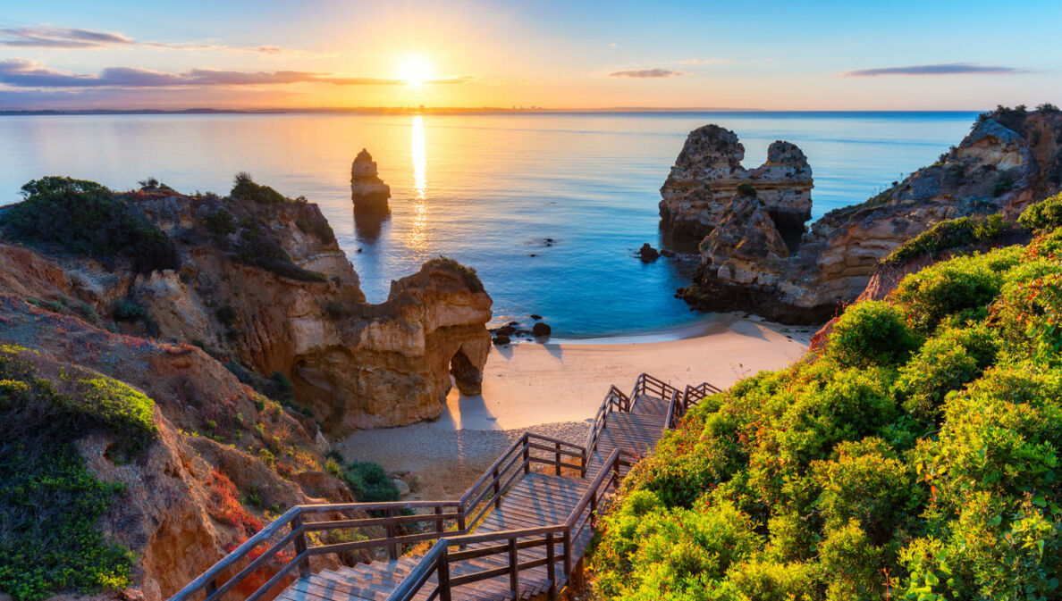 Der Strand Praia do Camilo in Portugal bei Sonnenuntergang, zu dem über Felsen eine Holztreppe nach unten führt.