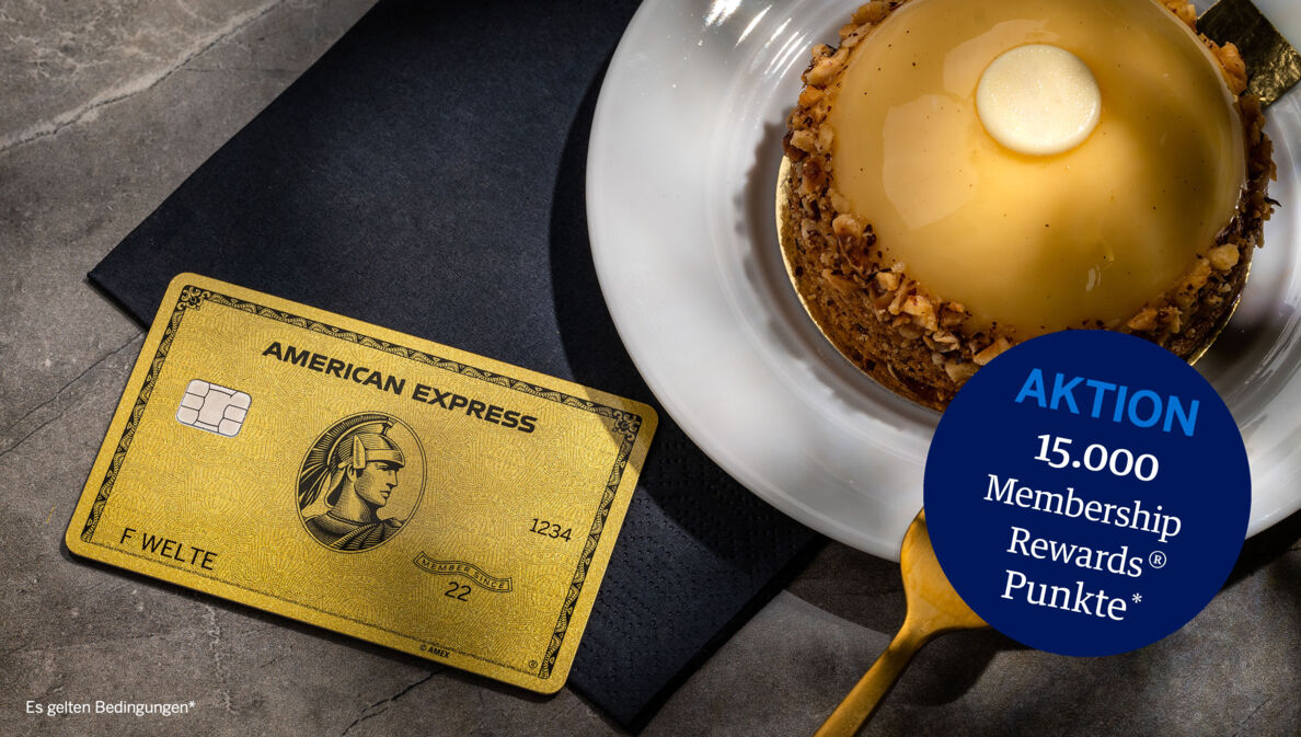 Eine goldene Kreditkarte von American Express liegt auf einer schwarzen Serviette neben einem goldfarbenen Törtchen auf einem Teller und einer goldenen Gabel.