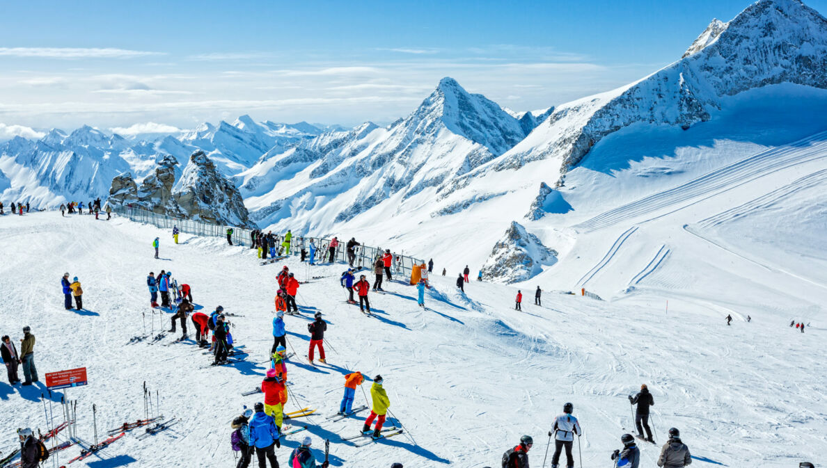 Skifahrende auf einem Plateau auf einer Skipiste vor schneebedecktem Gipfelpanorama.