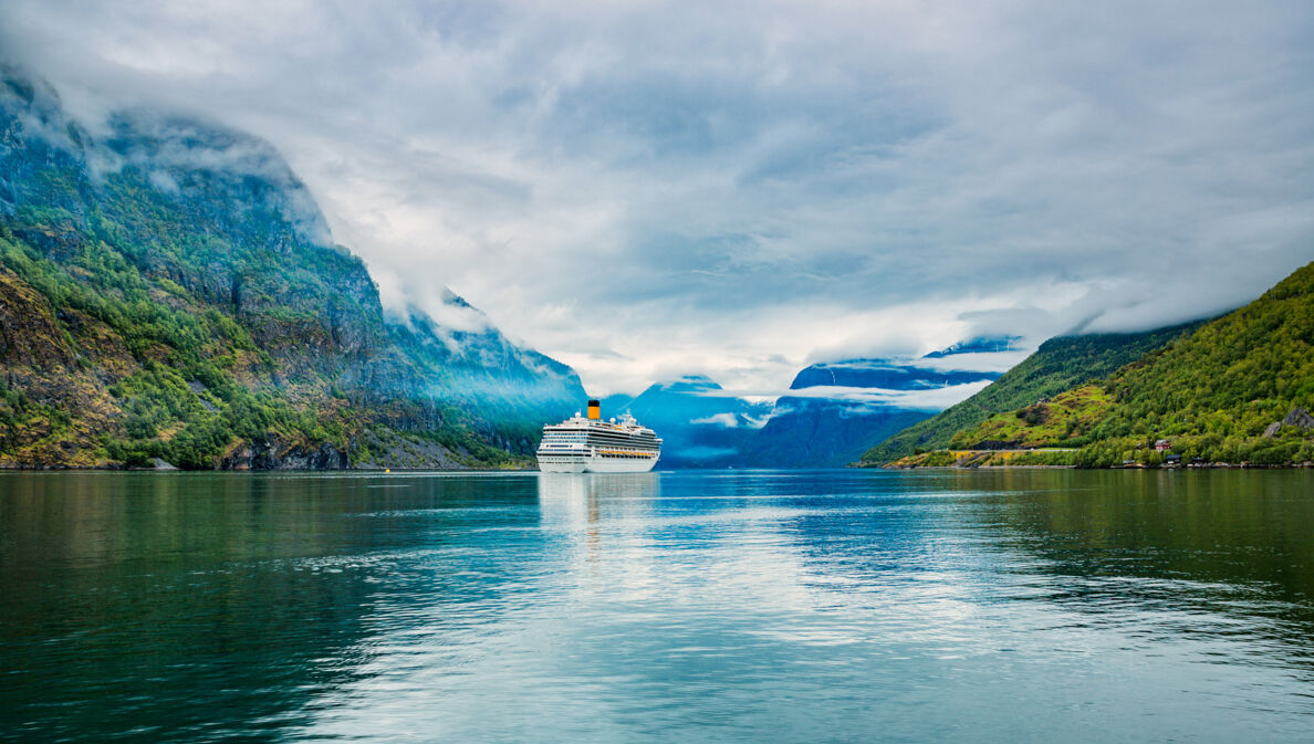 Kreuzfahrtschiff in wolkenverhangener Fjordlandschaft.