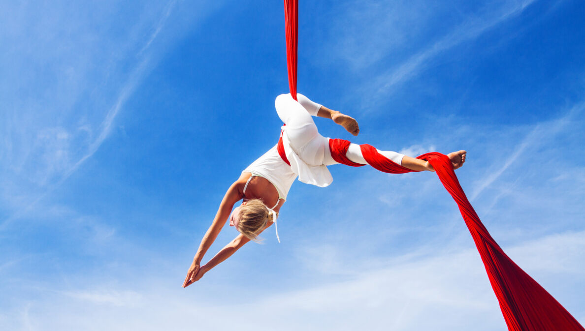 Untersicht einer Frau in weißer Kleidung in schwebender Yoga-Pose in einem roten Tuch vor blauem Himmel.